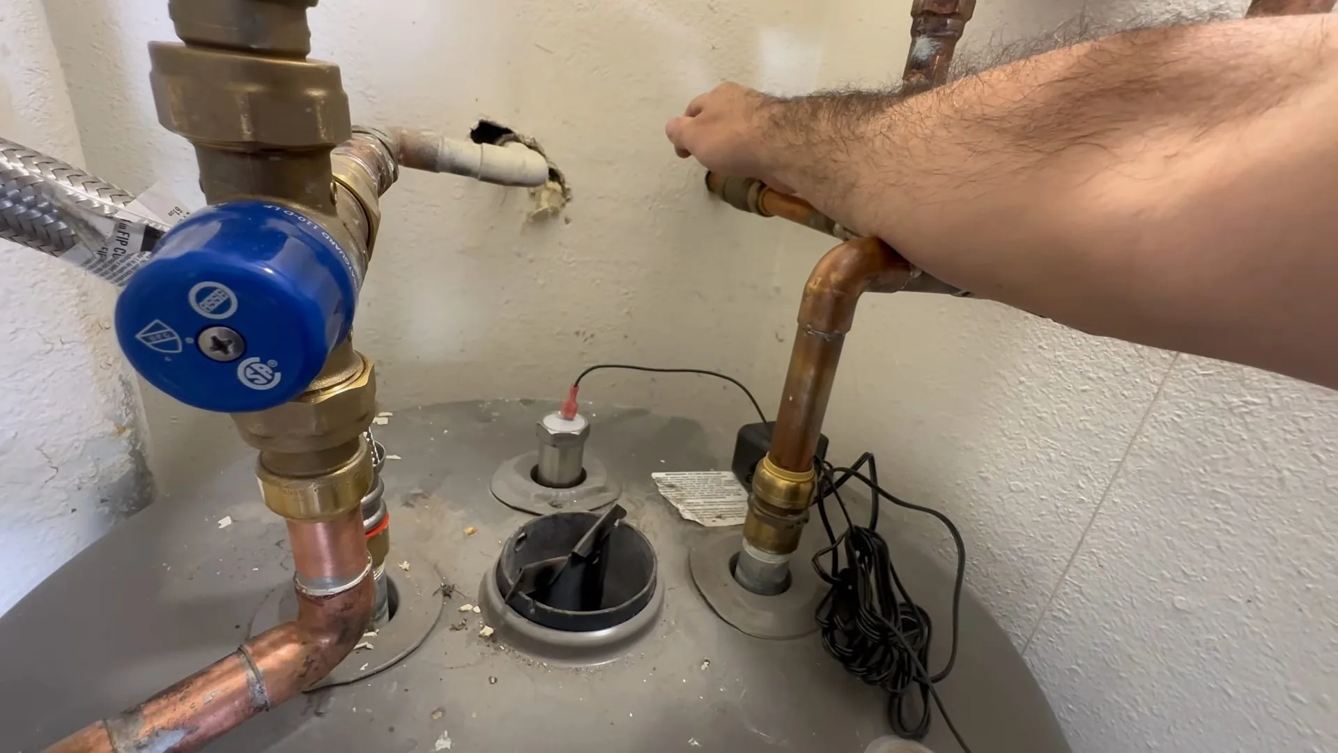 Arm reaching toward copper lines and a blue handled valve above the gray top of a tank water heater