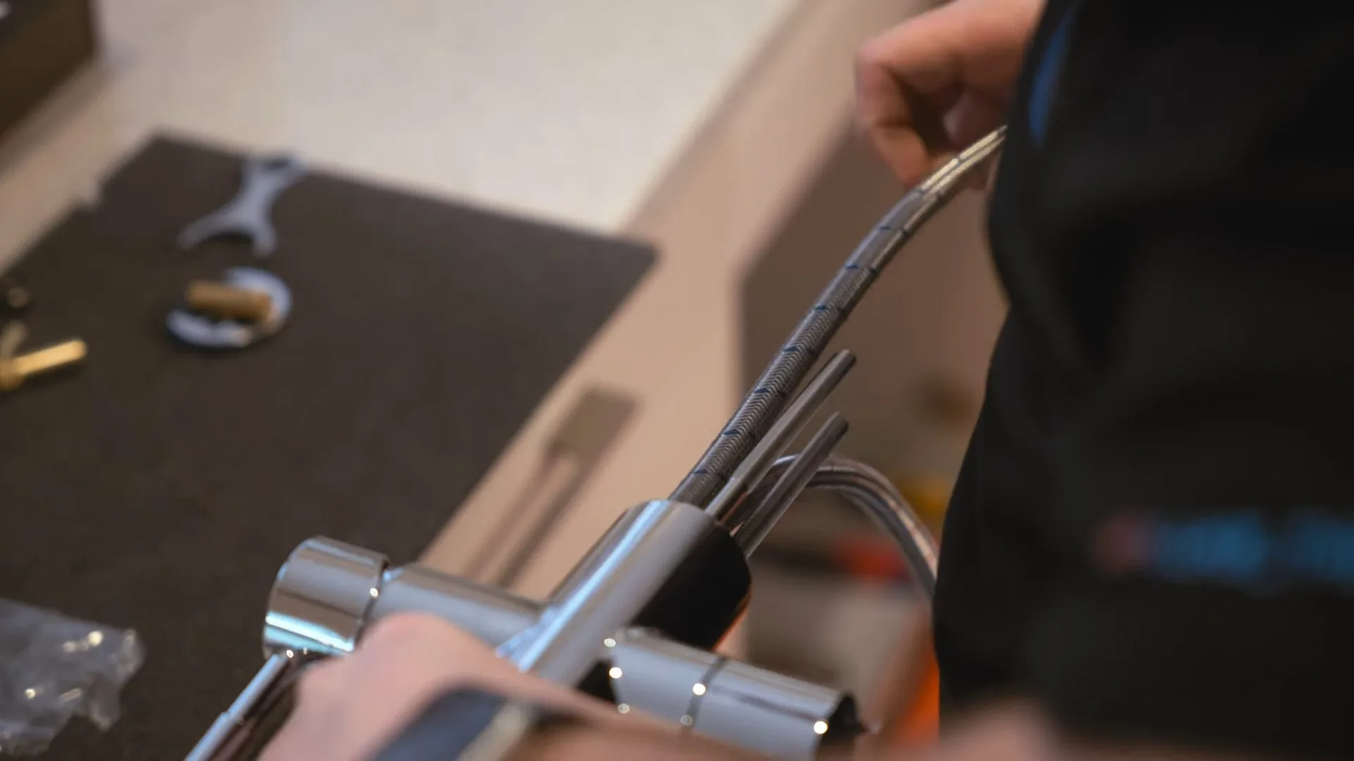Hands guiding a braided hose from a chrome faucet laid on a counter next to wrenches and brass fittings