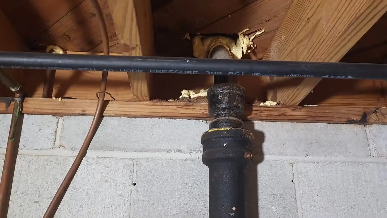 Black cast iron drain section meeting a plastic line beneath floor joists above a concrete block wall