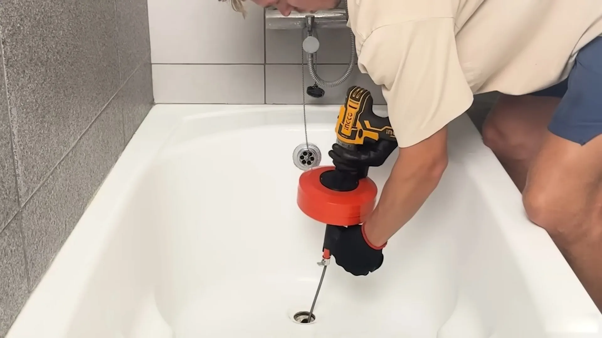 Person leaning into a bathtub and feeding a drill powered drain auger into the tub drain