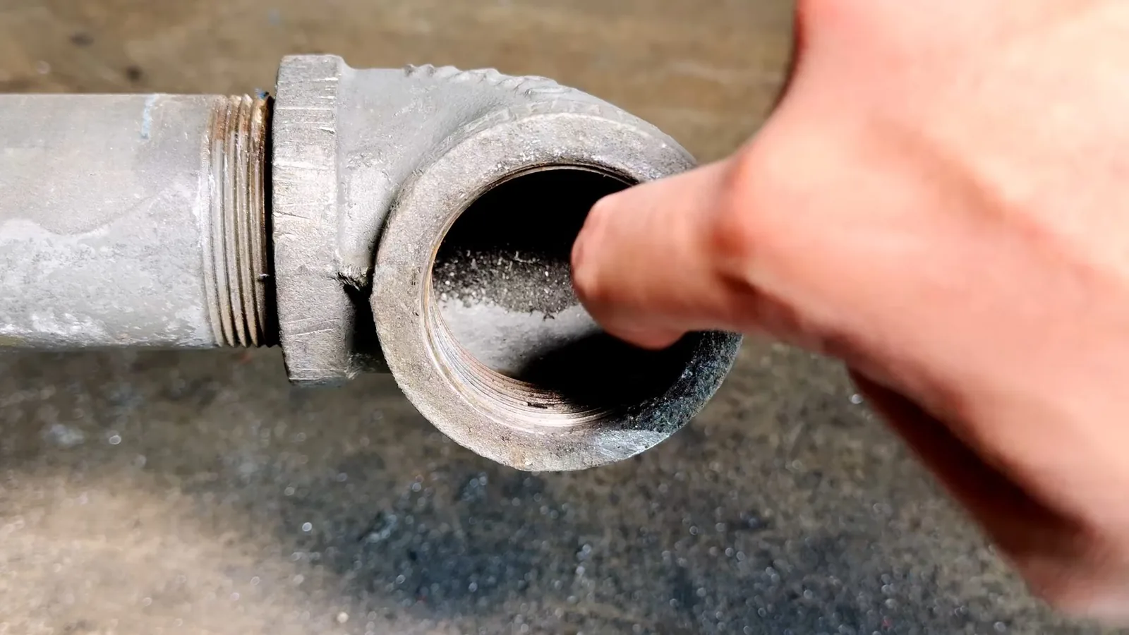 Finger pointing into the open end of a galvanized drain elbow with dark residue on the inside wall