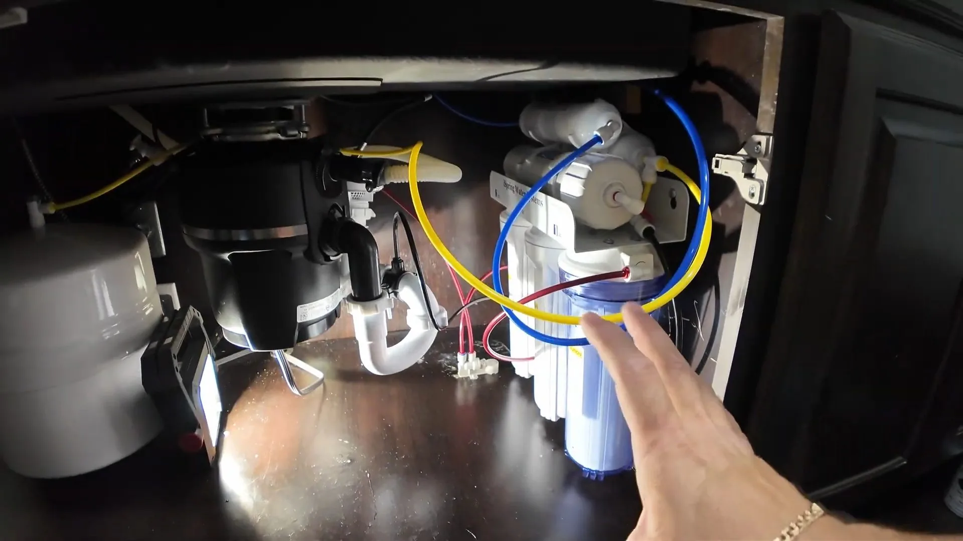 Black garbage disposal mounted under a sink with a white trap, blue and yellow tubing and filter housings inside a dark cabinet
