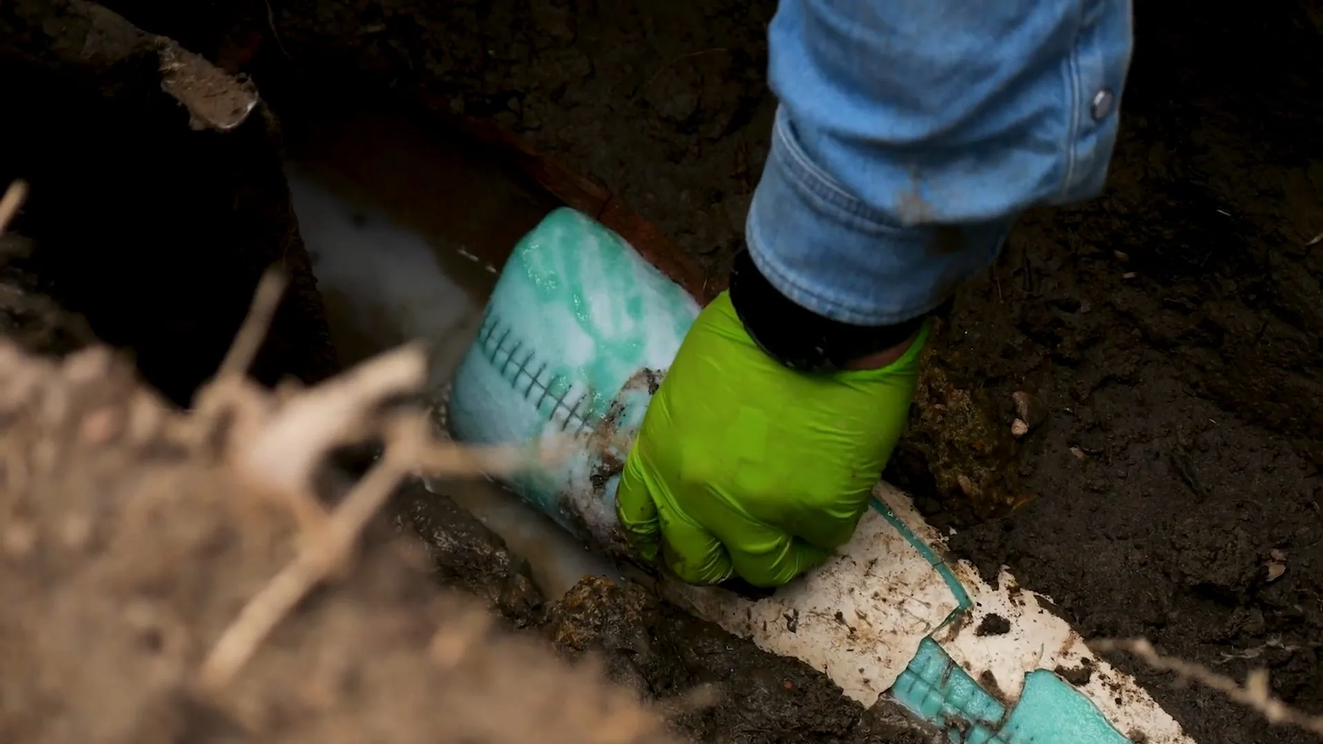 Green gloved hand handling a white and teal liner tube on soil in an excavation