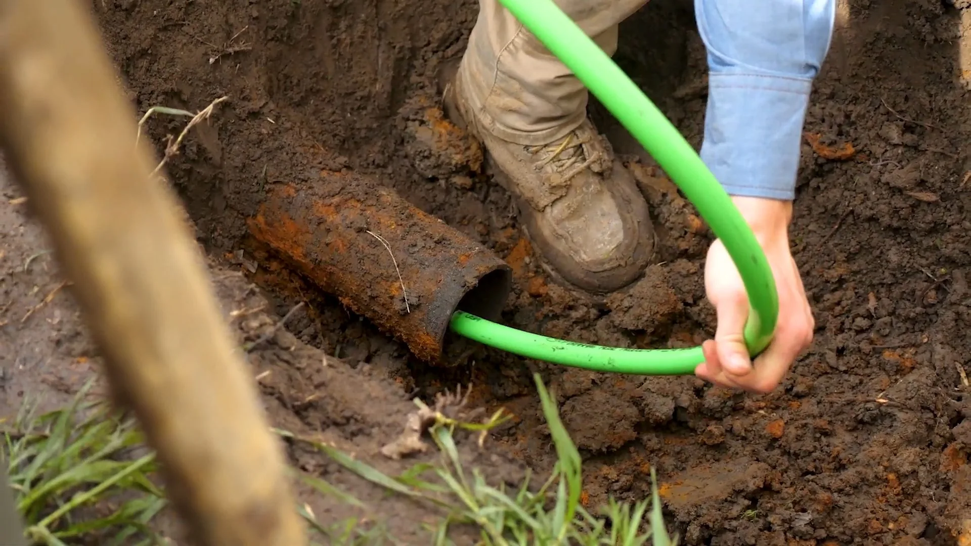Gloved hand guiding a green hose into the open end of a rusty pipe exposed in a dirt trench