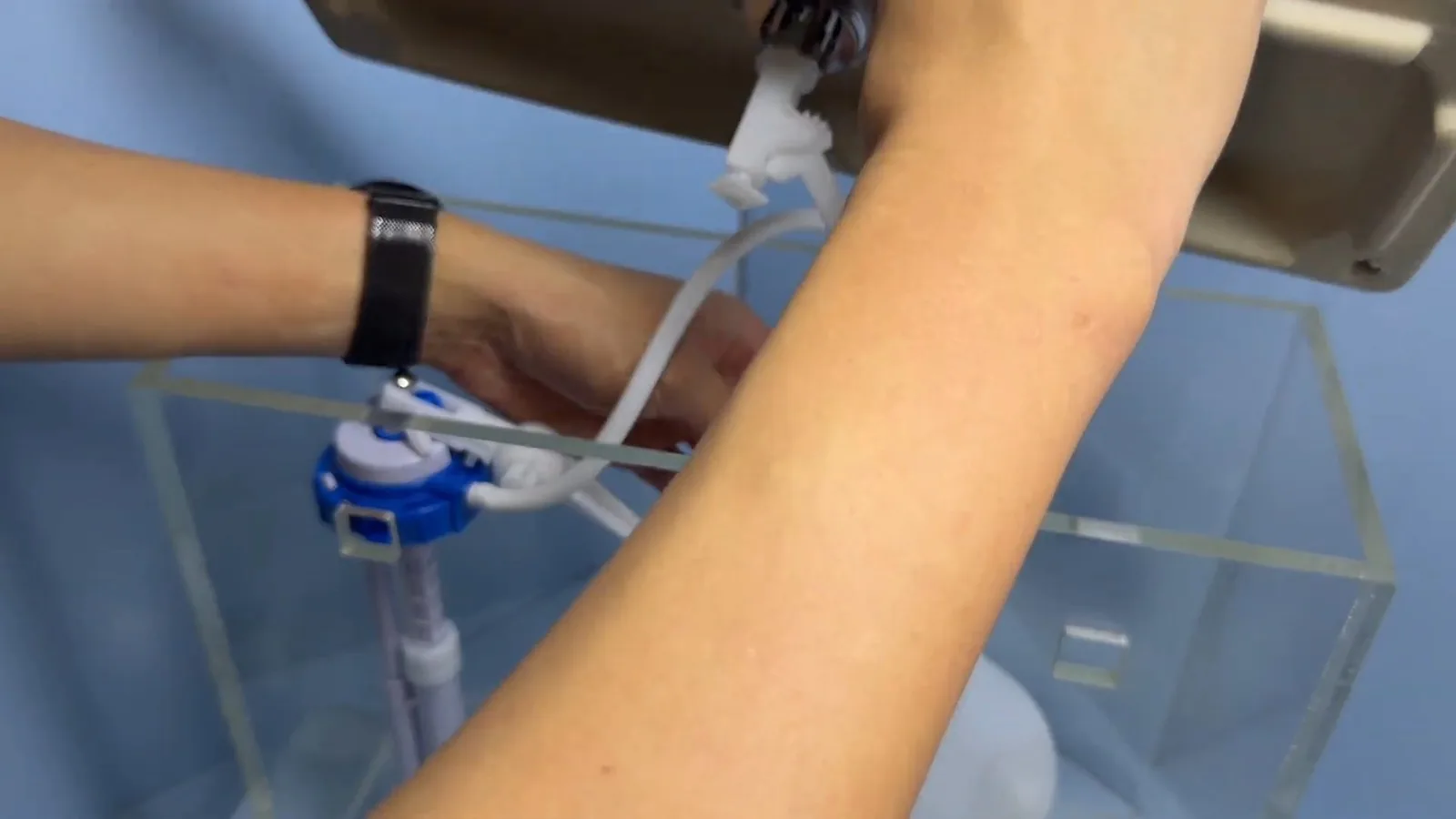 Hand adjusting a blue and white fill valve inside a clear toilet tank