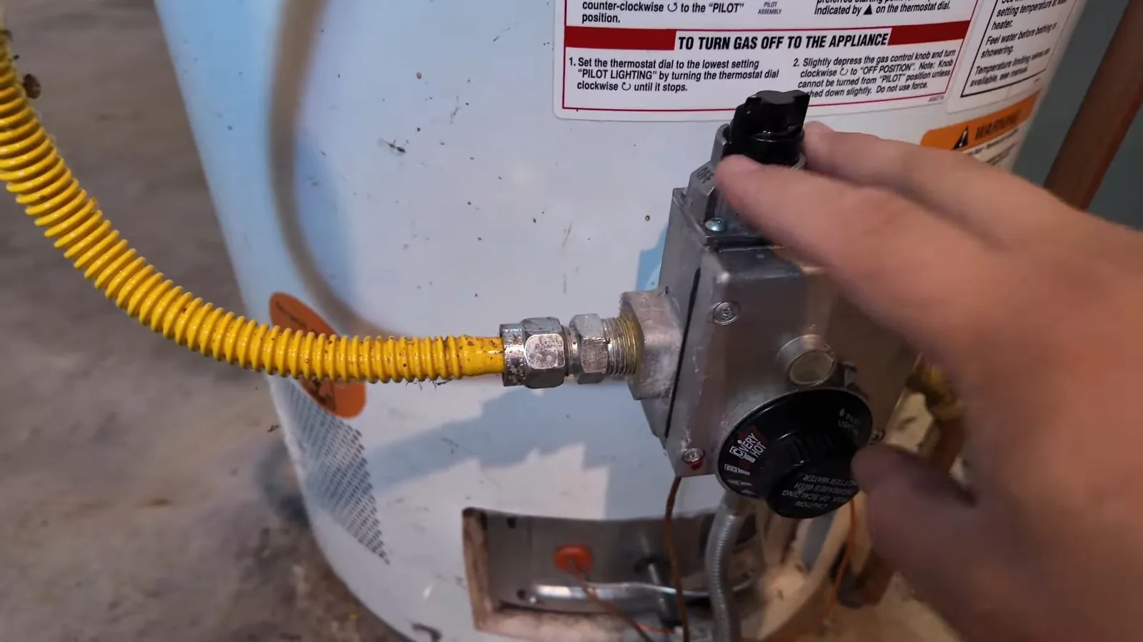 Hand adjusting the gas control knob on a tank water heater beside a yellow flexible gas line