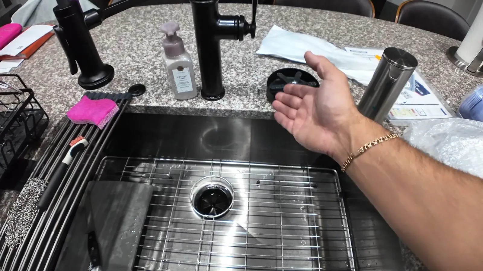 Open hand gesturing over a new stainless steel sink with a drain strainer, beside black faucets and unopened packaging