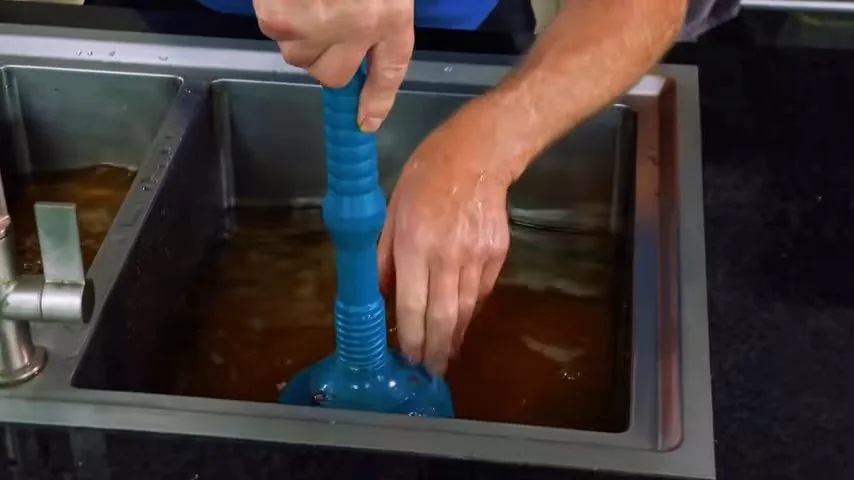 Hand pushing a blue plunger down into a stainless sink filled with brown standing water