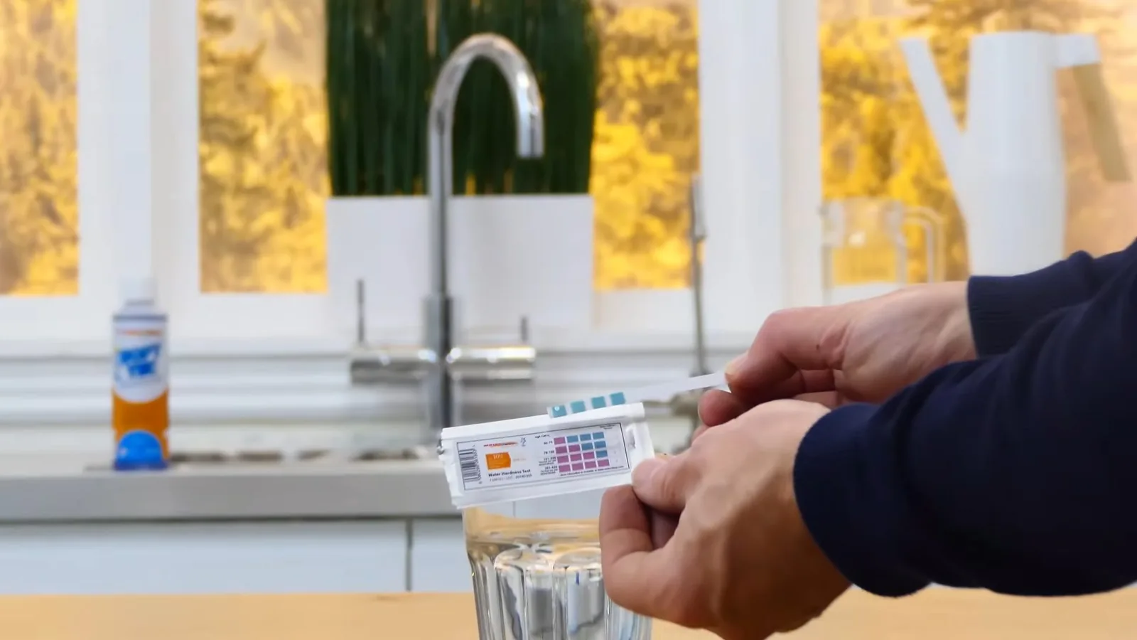 Hands holding a water hardness test strip over a glass of water in front of a kitchen faucet