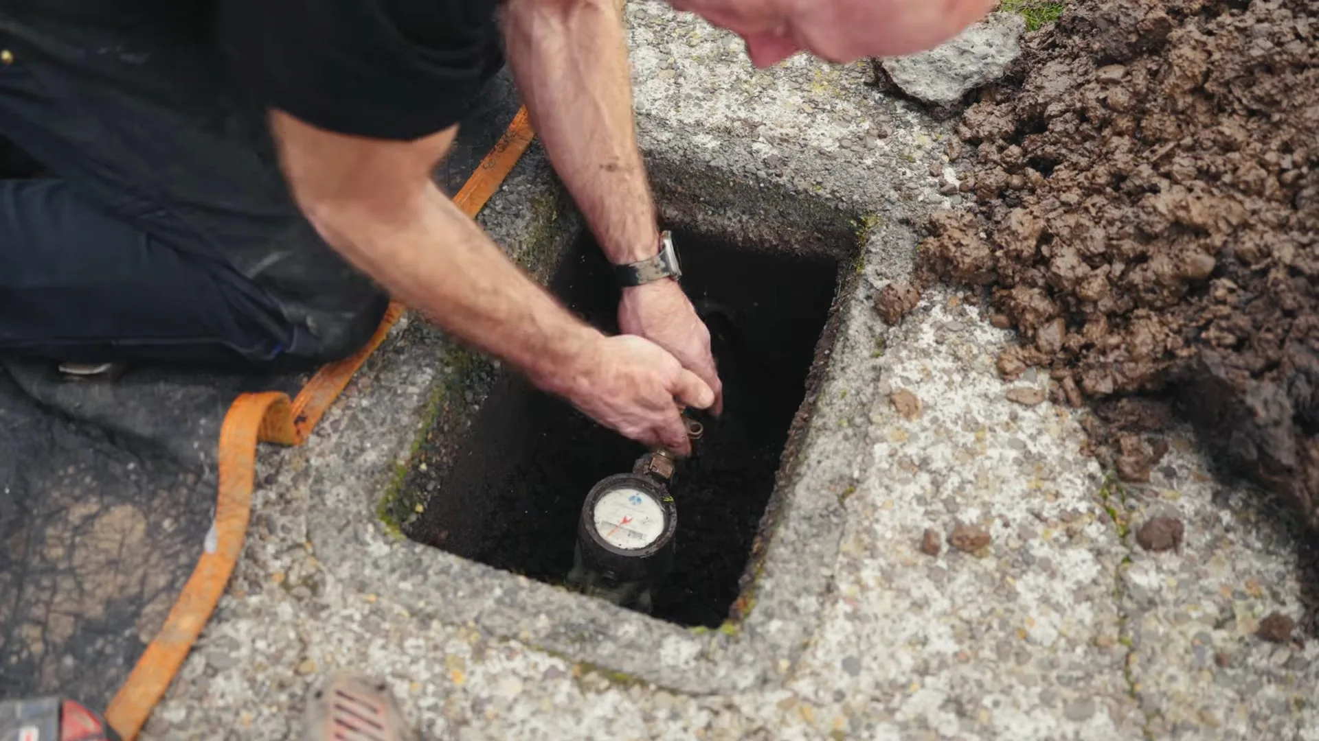 Man leaning over an open concrete box in the ground and reaching down to check the water meter inside
