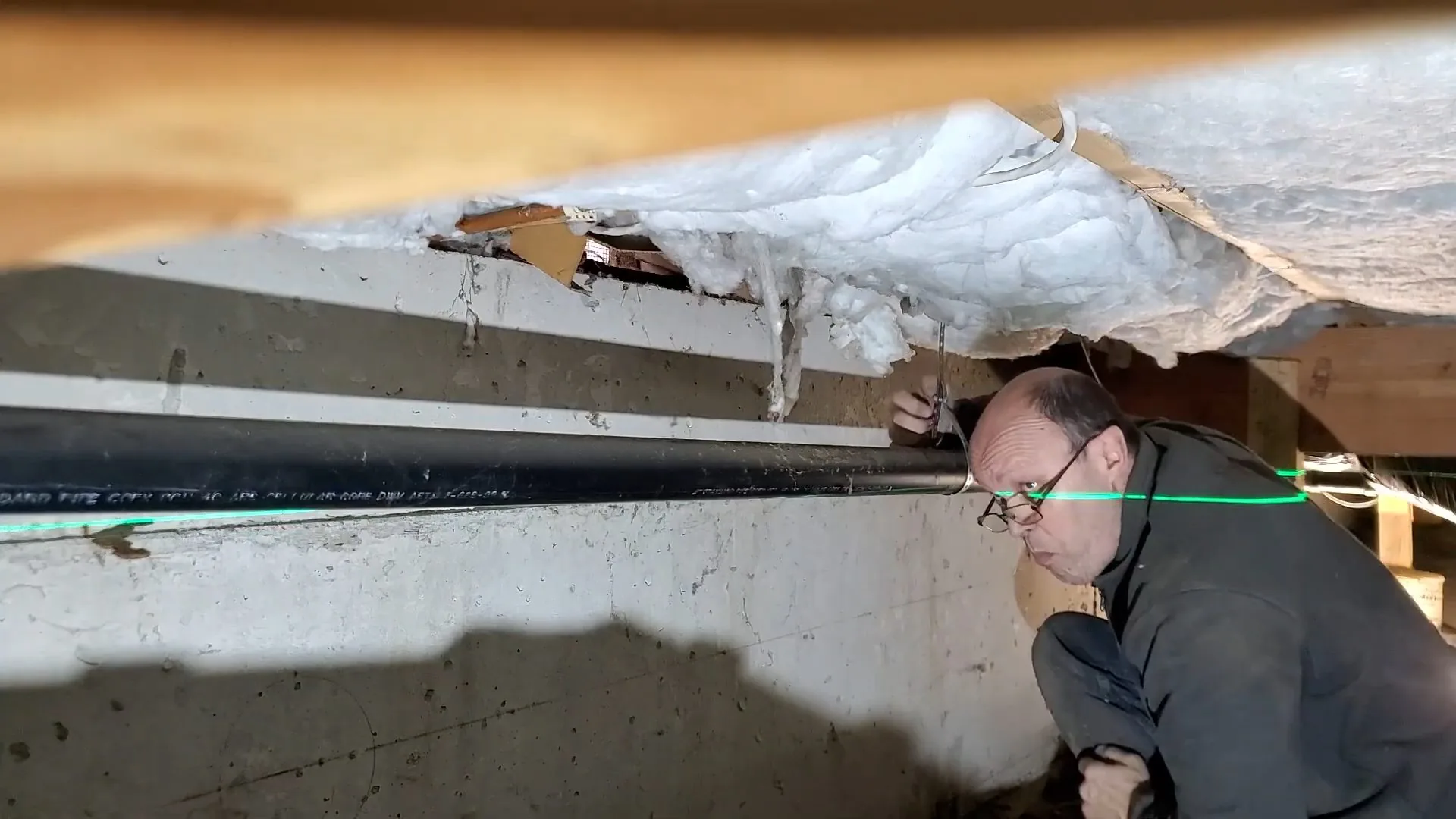 Man crouching in a crawlspace beside a long black pipe, looking under torn insulation with a green line laser