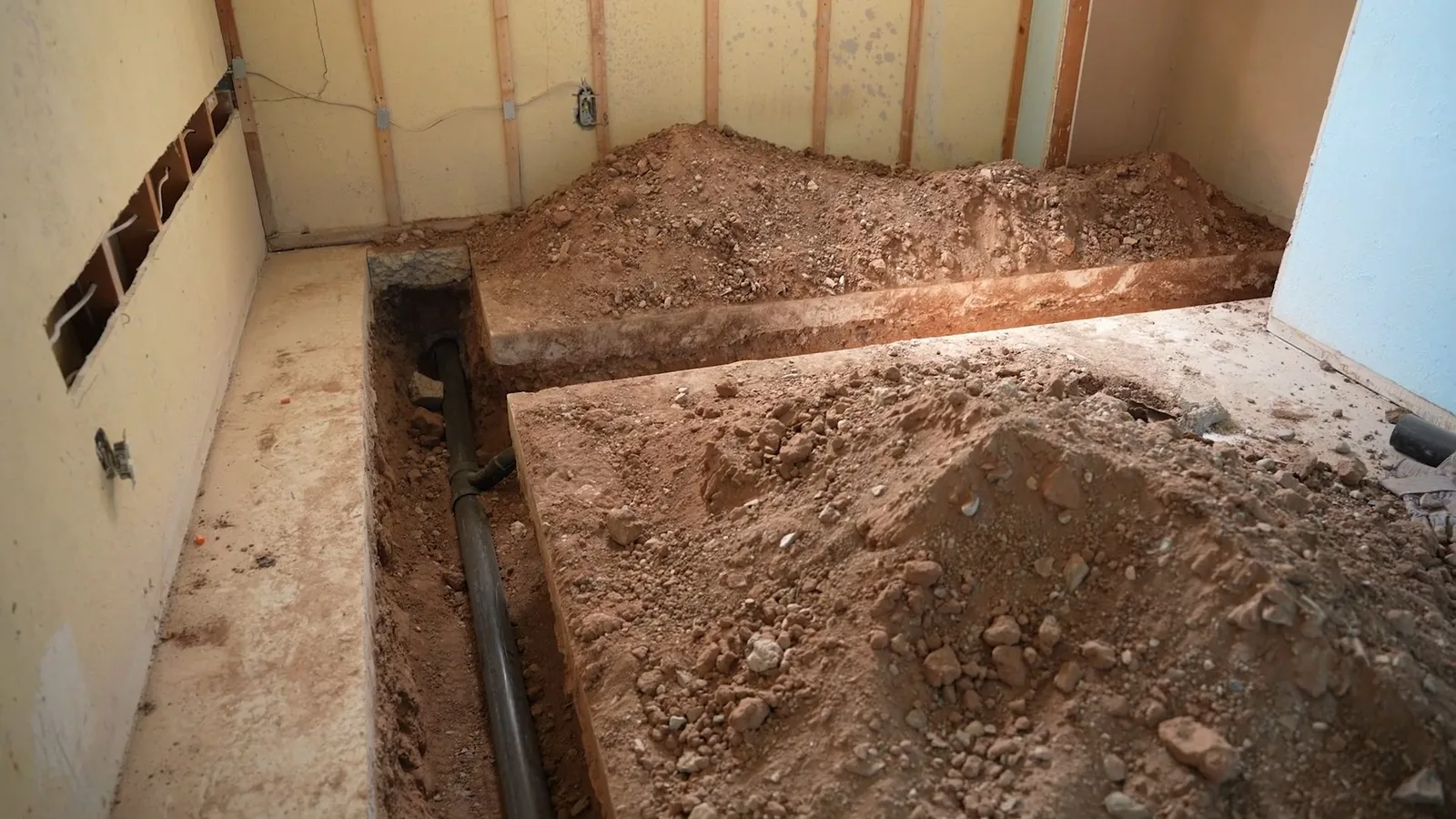 Narrow trench cut through a room floor with soil piled on both sides and a dark pipe exposed at the bottom