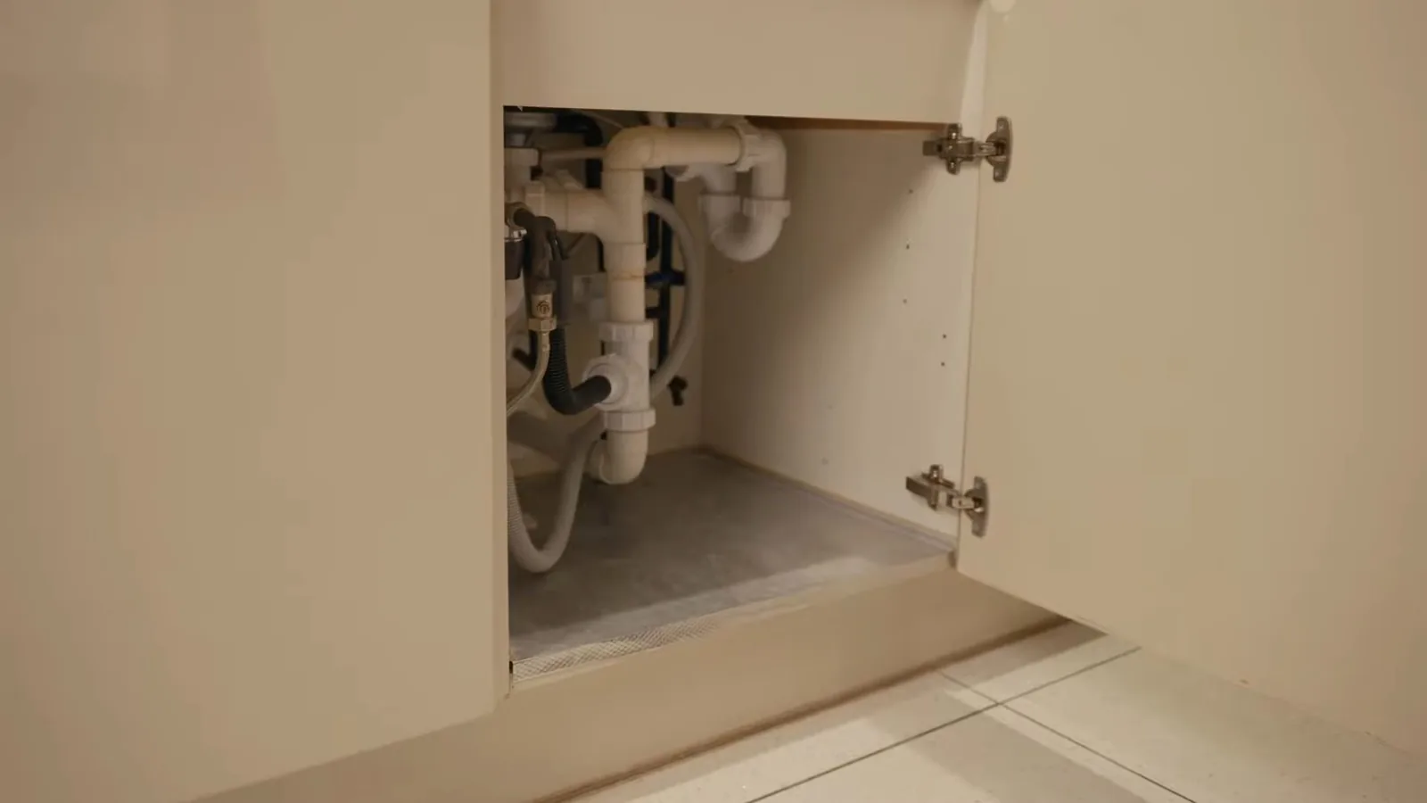 Open cabinet door showing white drain pipes, a trap and several hoses in the space under a sink