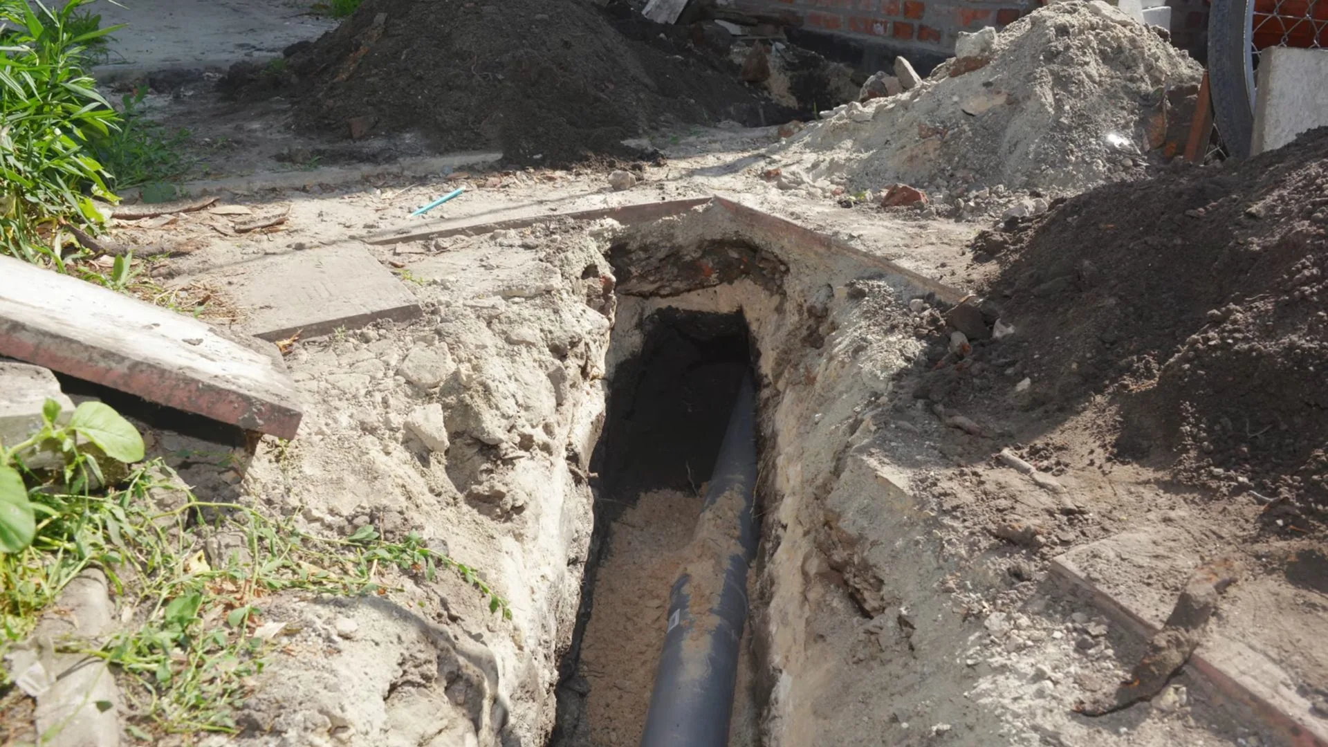 Open trench in a yard with a dark drain pipe exposed at the bottom between piles of soil