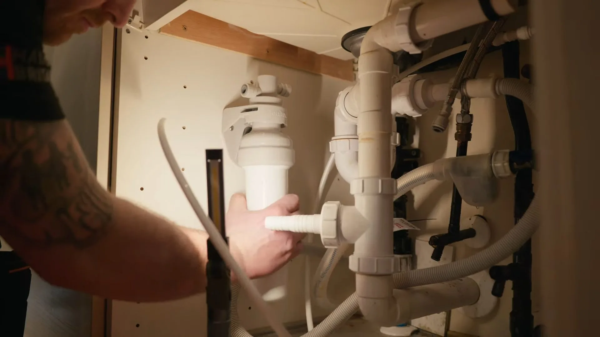 Tattooed forearm and hand holding a white filter housing in a crowded cabinet under a kitchen sink