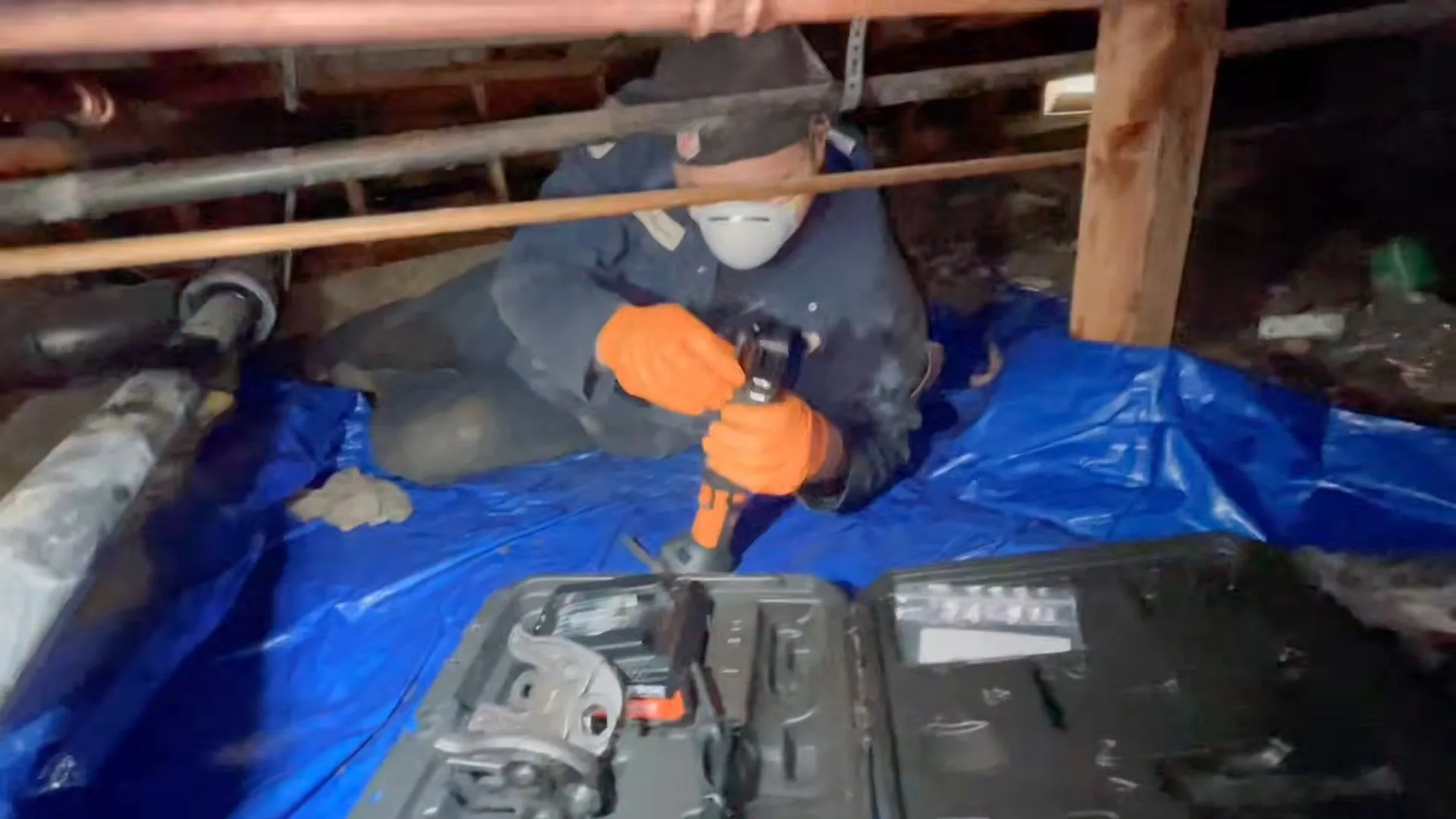 Plumber in a respirator mask kneeling on a blue tarp in a crawlspace and working a pipe tool above an open tool case
