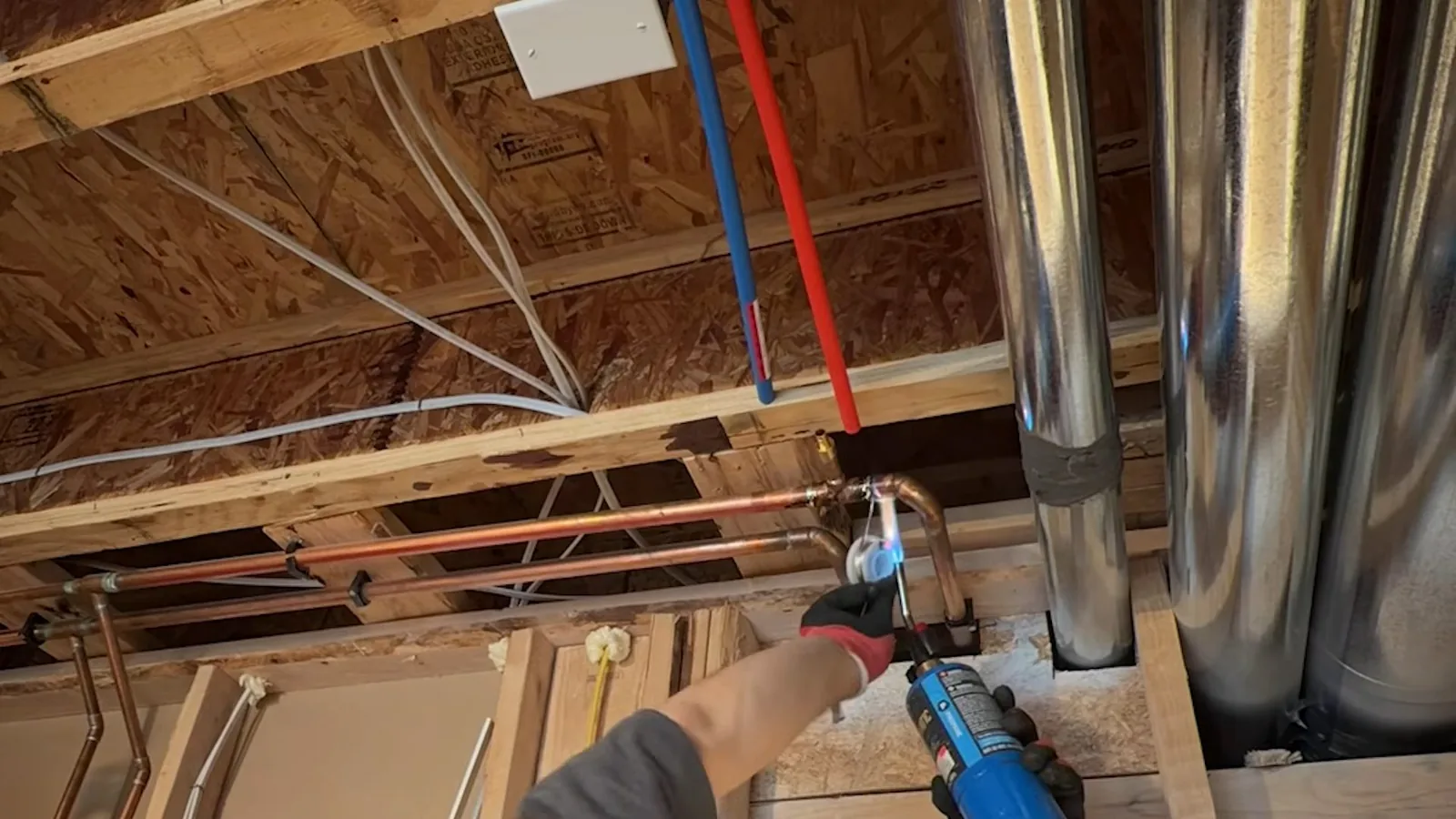 Plumber holding a torch to a copper joint among open floor joists with red and blue lines running overhead