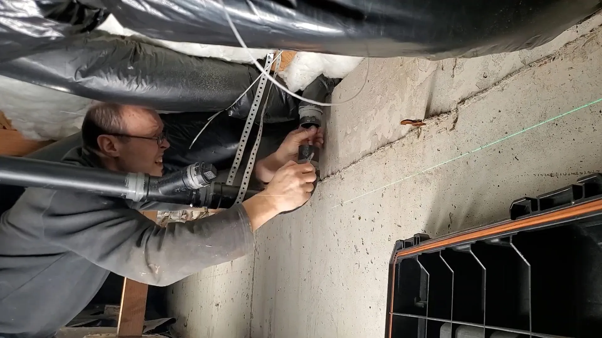 Plumber working on a drain pipe along a concrete foundation wall with ductwork overhead