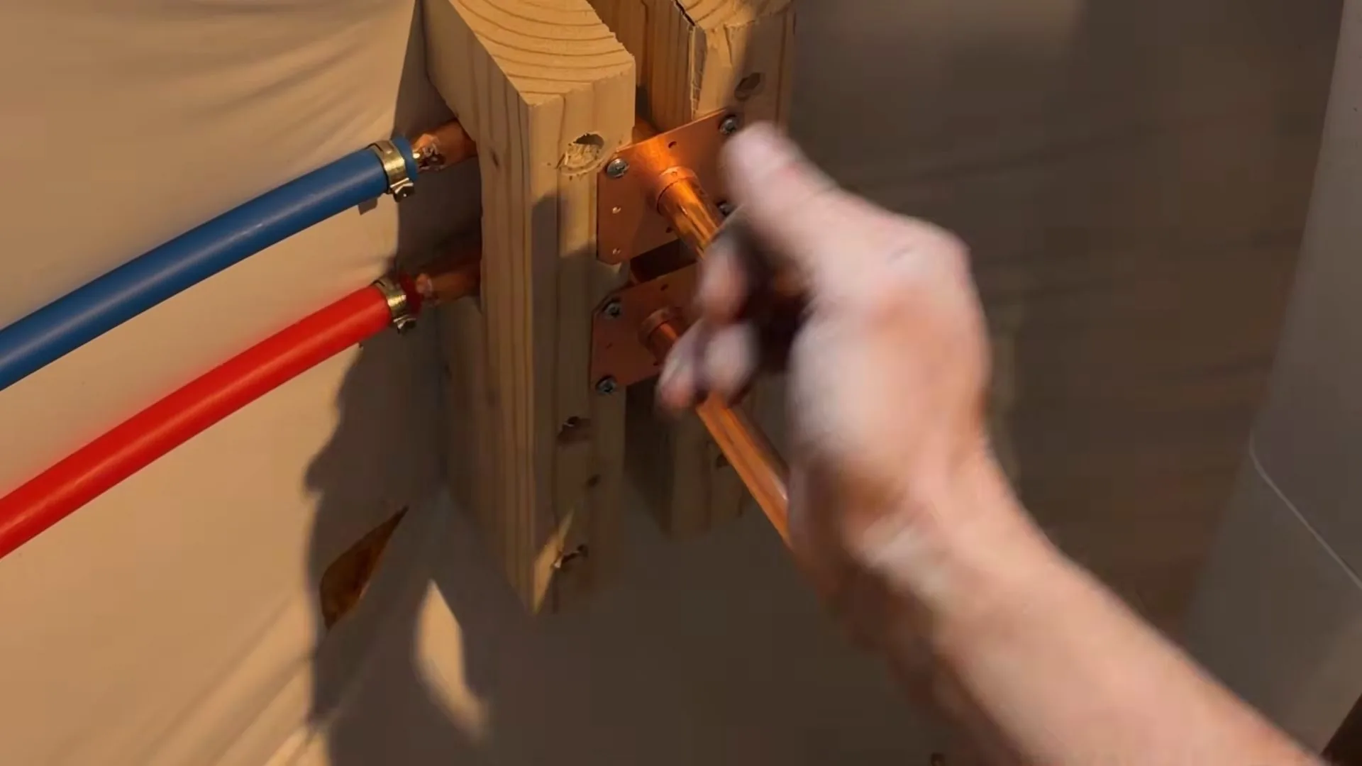 Hand steadying a copper stub where red and blue flexible supply lines connect on a wood mounting board