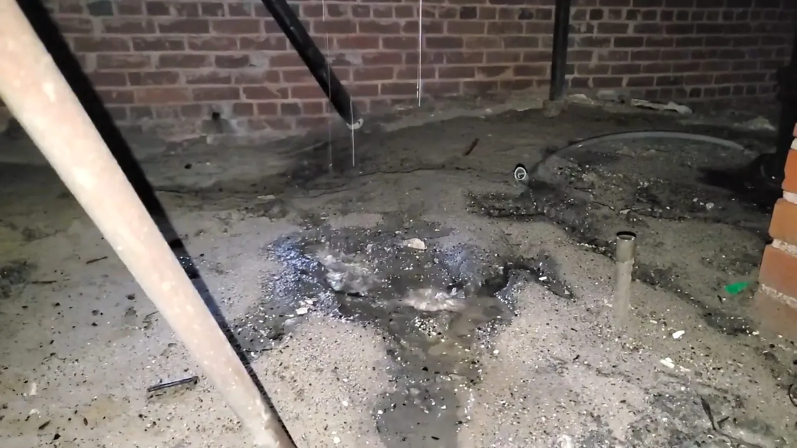 Water dripping from a pipe onto a wet, muddy crawlspace floor beside a brick foundation wall