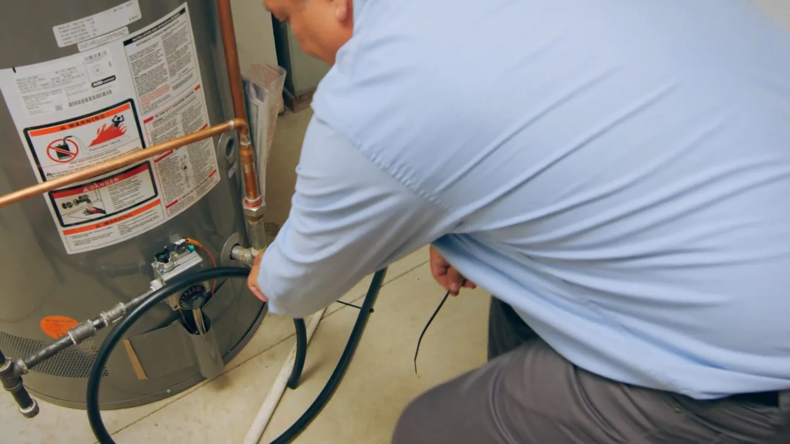 Technician in a light blue shirt kneeling beside a gas water heater and working at a hose near its base