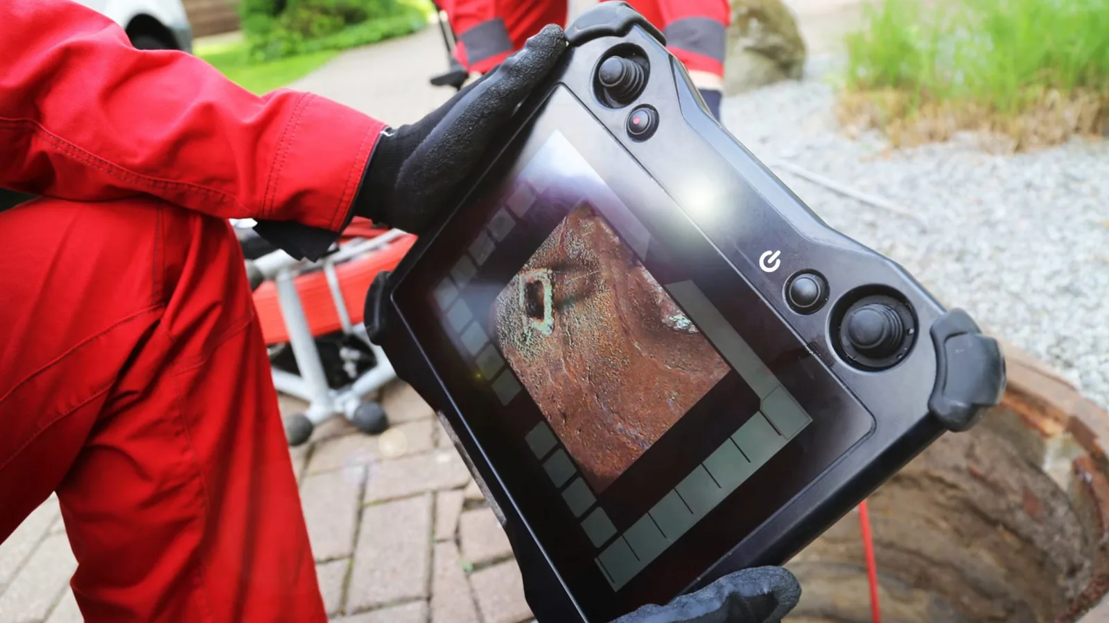 Technician in red coveralls holding a handheld drain camera monitor that shows the inside of a pipe