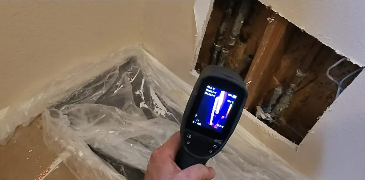 Handheld thermal camera aimed at an opened wall corner where plastic sheeting covers the floor