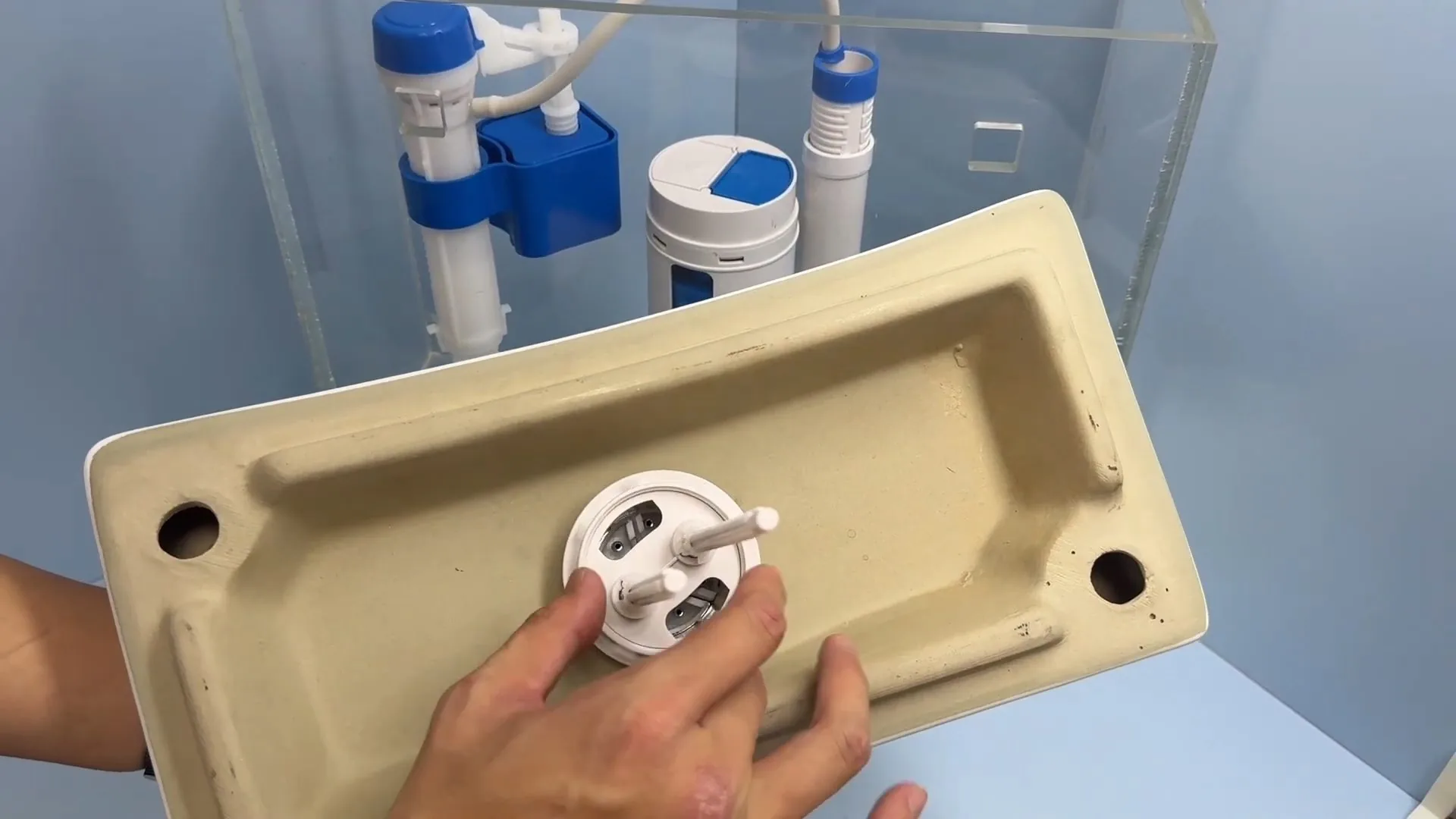 Ceramic toilet tank lid held upside down beside a clear tank showing the fill valve and flush valve