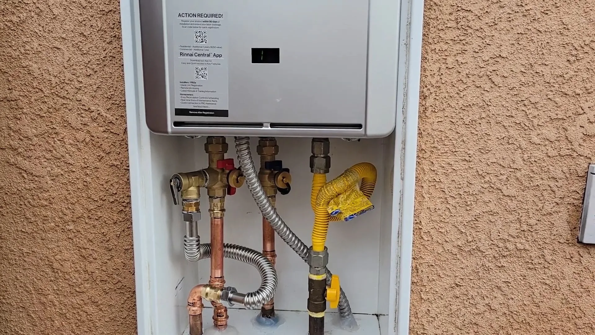 Wall mounted tankless water heater in an outdoor alcove above copper water lines, shutoff valves and a yellow gas connector